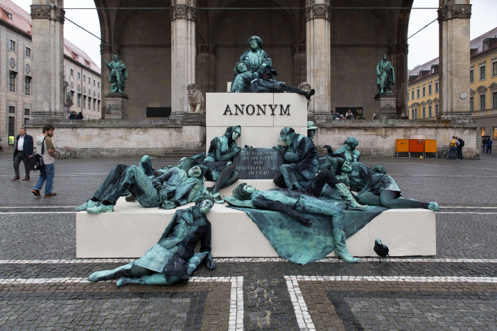 Photograph of an art project by Silke Wagner on Odeonsplatz in front of the Feldherrnhalle. The installation is a living monument intended to create the illusion of a sculpture: A series of performers sit and lie in motionless poses on white plinths stacked like pyramids. Their faces show rigid, sad or agonised expressions. The performers are dressed in green-grey costumes that match the verdigris bronze patina of the two generals' sculptures on the left and right in the Feldherrnhalle. Two of the figures in the foreground hold a memorial plaque with the inscription "In memory of the victims of sexual violence during the war". The highest plinth block also bears the word "Anonymous" in capital letters, which is also the title of the art project. People stand around the living monument and look at the installation.