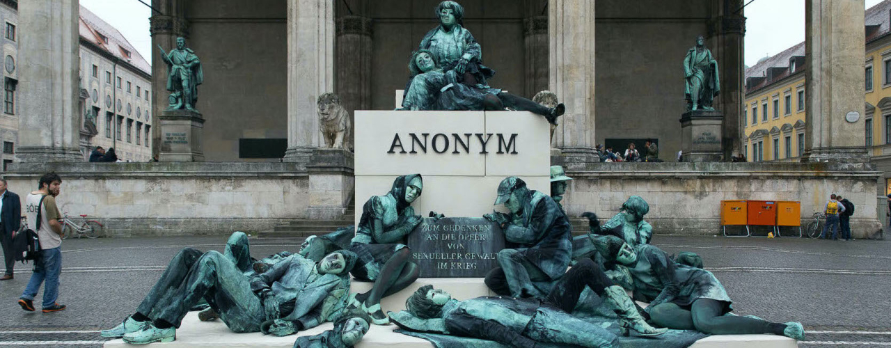 Photograph of an art project by Silke Wagner on Odeonsplatz in front of the Feldherrnhalle. The installation is a living monument intended to create the illusion of a sculpture: A series of performers sit and lie in motionless poses on white plinths stacked like pyramids. Their faces show rigid, sad or agonised expressions. The performers are dressed in green-grey costumes that match the verdigris bronze patina of the two generals' sculptures on the left and right in the Feldherrnhalle. Two of the figures in the foreground hold a memorial plaque with the inscription "In memory of the victims of sexual violence during the war". The highest plinth block also bears the word "Anonymous" in capital letters, which is also the title of the art project.