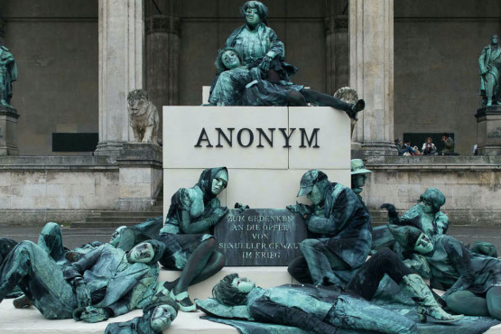 Photograph of an art project by Silke Wagner on Odeonsplatz in front of the Feldherrnhalle. The installation is a living monument intended to create the illusion of a sculpture: A series of performers sit and lie in motionless poses on white plinths stacked like pyramids. Their faces show rigid, sad or agonised expressions. The performers are dressed in green-grey costumes that match the verdigris bronze patina of the two generals' sculptures on the left and right in the Feldherrnhalle. Two of the figures in the foreground hold a memorial plaque with the inscription "In memory of the victims of sexual violence during the war". The highest plinth block also bears the word "Anonymous" in capital letters, which is also the title of the art project.