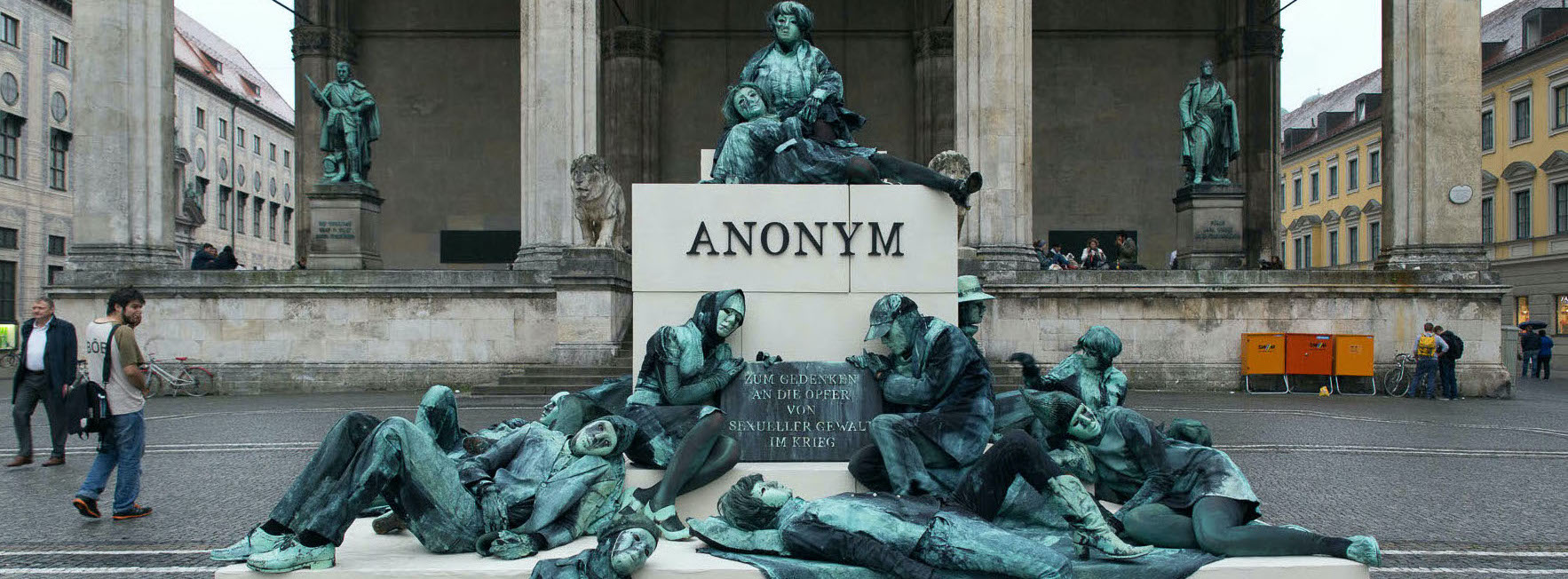 Photograph of an art project by Silke Wagner on Odeonsplatz in front of the Feldherrnhalle. The installation is a living monument intended to create the illusion of a sculpture: A series of performers sit and lie in motionless poses on white plinths stacked like pyramids. Their faces show rigid, sad or agonised expressions. The performers are dressed in green-grey costumes that match the verdigris bronze patina of the two generals' sculptures on the left and right in the Feldherrnhalle. Two of the figures in the foreground hold a memorial plaque with the inscription "In memory of the victims of sexual violence during the war". The highest plinth block also bears the word "Anonymous" in capital letters, which is also the title of the art project.