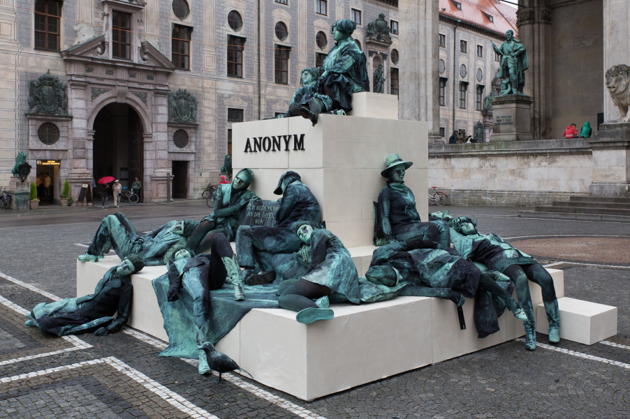 Side view of an art project by Silke Wagner on the Odeonsplatz in front of the Feldherrnhalle. The installation is a living monument designed to create the illusion of a sculpture: A series of performers sit and lie in motionless poses on white pedestals stacked like pyramids. Their faces show expressions of rigidity, sadness or torture. The performers are dressed in green-grey costumes that match the verdigris bronze patina of the commander sculptures of the Feldherrnhalle. Two of the figures in the foreground hold a plaque with the inscription 'In memory of the victims of sexual violence during the war'. The word "Anonymous", which is also the title of the art project, is written in capital letters on the highest plinth.