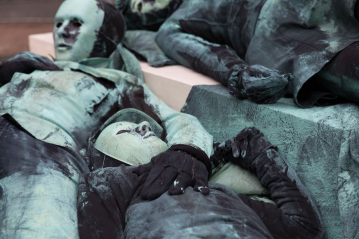 Close-up of an art project by Silke Wagner on Odeonsplatz. The installation is a living monument intended to create the illusion of a sculpture: The photograph shows three performers lying on two white pedestals stacked like pyramids. The people are wearing masks and headgear. The performers are wearing green-grey costumes reminiscent of the verdigris bronze patina of the generals' sculptures in the Feldherrnhalle.