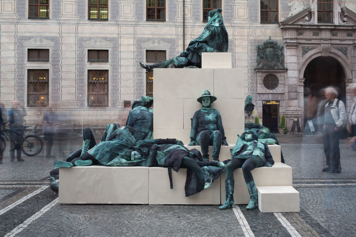 Seitliche Fotografie eines Kunstprojektes von Silke Wagner am Odeonsplatz vor der Feldherrnhalle. Die Installation ist ein lebendes Denkmal, das die Illusion einer Skulptur erzeugen soll: Auf pyramidenartig gestapelten weißen Sockeln sitzen und liegen in regungsloser Pose eine Reihe von Performer*innen. Ihre Gesichter zeigen starre, traurige oder auch gequälte Ausdrücke. Die Performer*innen sind in grün-graue Kostüme gekleidet, die sich der Grünspan-Bronzepatina der Feldherrenskulpturen in der Feldherrnhalle anpassen.