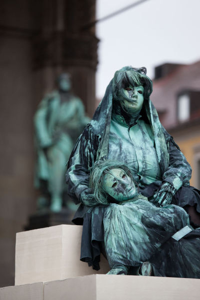 Close-up of an art project by Silke Wagner on Odeonsplatz in front of the Feldherrnhalle. The installation is a living monument designed to create the illusion of a sculpture: Two female performers can be seen in a motionless pose on white plinths stacked like pyramids. One woman sits on the upper plinth, wearing a veil over her hair. The second woman on the lower plinth is leaning on her arms, her head resting in the lap of the other woman. The two performers wear green-grey costumes reminiscent of the verdigris bronze patina of the generals' sculptures in the Feldherrnhalle.