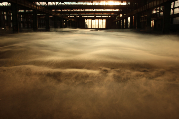 Blick in die leere Industriehalle 24 des ehemaligen Reichsbahn-Ausbesserungswerk Freimann bei Sonnenuntergang. Die Sonne fällt durch großen Glasfenster. Durch die Halle wabern dicke Nebelschwaden.