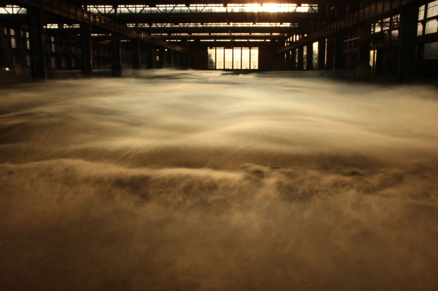 View into the empty industrial hall 24 of the former Freimann railway repair works at sunset. The sun shines through large glass windows. Thick wafts of mist waft through the hall.