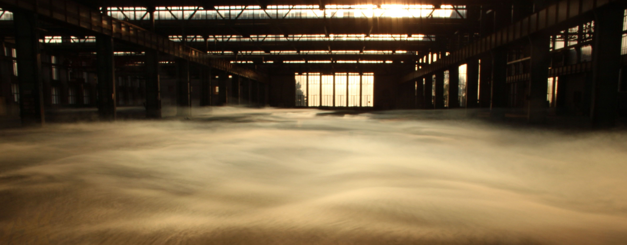 View into the empty industrial hall 24 of the former Freimann railway repair works at sunset. The sun shines through large glass windows. Thick wafts of mist waft through the hall.