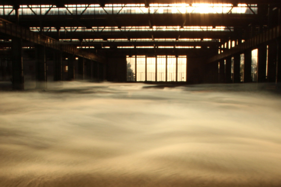 View into the empty industrial hall 24 of the former Freimann railway repair works at sunset. The sun shines through large glass windows. Thick wafts of mist waft through the hall.