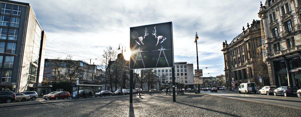 View of the billboard on Lenbachplatz looking towards the city centre with the motif "Homo Alpinus" by Stefano Giuriati. The motif shows on black background a mirror image of the artist Stefano Giuriati in a wrestling jersey with a deer antler on his head standing on a ladder. He is giving the impression that he is entering into a fight with himself.