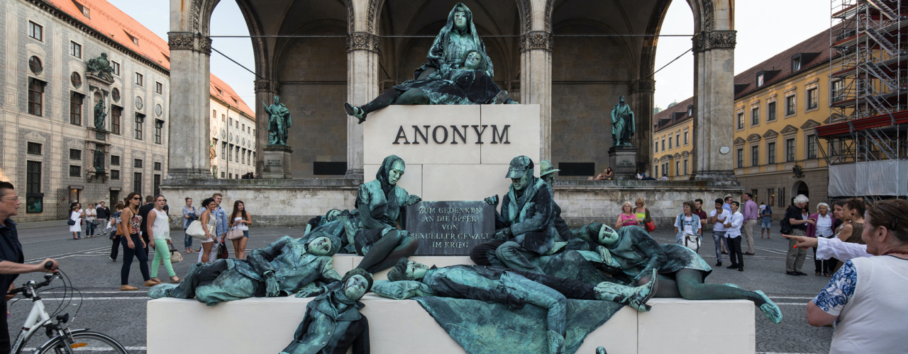 Photograph of an art project by Silke Wagner on Odeonsplatz in front of the Feldherrnhalle. The installation is a living monument intended to create the illusion of a sculpture: A series of performers sit and lie in motionless poses on white plinths stacked like pyramids. Their faces show rigid, sad or agonised expressions. The performers are dressed in green-grey costumes that match the verdigris bronze patina of the two generals' sculptures on the left and right in the Feldherrnhalle. Two of the figures in the foreground hold a memorial plaque with the inscription "In memory of the victims of sexual violence during the war". The highest plinth block also bears the word "Anonymous" in capital letters, which is also the title of the art project.