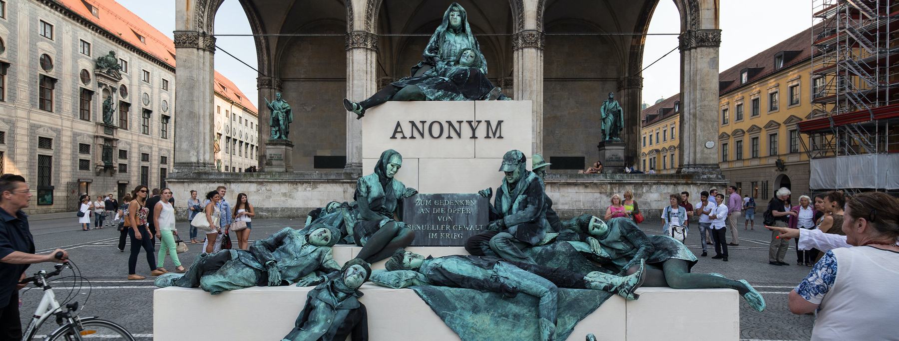 Photograph of an art project by Silke Wagner on Odeonsplatz in front of the Feldherrnhalle. The installation is a living monument intended to create the illusion of a sculpture: A series of performers sit and lie in motionless poses on white plinths stacked like pyramids. Their faces show rigid, sad or agonised expressions. The performers are dressed in green-grey costumes that match the verdigris bronze patina of the two generals' sculptures on the left and right in the Feldherrnhalle. Two of the figures in the foreground hold a memorial plaque with the inscription "In memory of the victims of sexual violence during the war". The highest plinth block also bears the word "Anonymous" in capital letters, which is also the title of the art project.