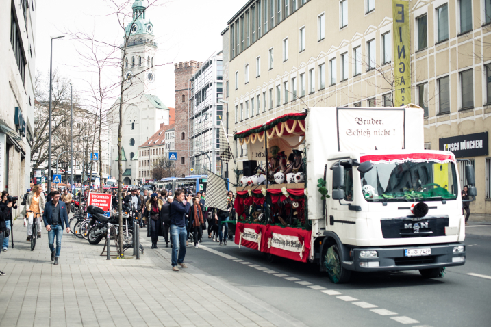 Fotografie eines Demonstrationszug auf der Straße am Oberanger in der Münchner Innenstadt. Der Zug wird von einem weißen dekorierten Festwagen angeführt, auf den Gruppen von Prozessionsteilnehmer*innen folgen. Der Festwagen ist mit roten Stoffen sowie weißen und grünen Girlanden und Papierblumen geschmückt. An der Frontseite des Lastwagens ist in altdeutscher Schrift ein Schild mit dem Text "Bruder Schieß nicht!" angebracht.