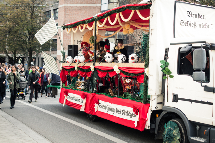 Foto einer Parade auf der Straße im Zentrum von München. Die Prozession wird von einem weiß geschmückten Wagen angeführt, gefolgt von Prozessionsteilnehmer*innen. Der Wagen ist mit rotem Stoff, weißen und grünen Girlanden und Papierblumen geschmückt. An ihm ist ein Schild mit der Aufschrift "Bruder Schieß nicht" in altdeutscher Sprache angebracht, und an der Seite hängt ein Transparent mit der Aufschrift "Demonstration zur Beerdigung des Krieges", ebenfalls in altdeutscher Sprache. Mehrere Frauen stehen auf dem Wagen und singen in Mikrofone, die an dem Wagen befestigt sind. Sie sind in farbenfrohe Trachten gekleidet, tragen Kopfbedeckungen und sind ausdrucksstark geschminkt.