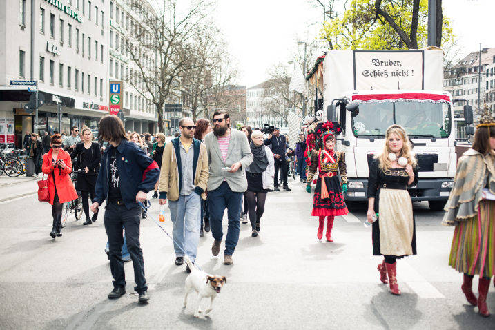 Foto eines Demonstrationszuges auf der mehrspurigen Straße am Karlsplatz in der Münchner Innenstadt. Der Festzug wird von einem Auto begleitet: Bei dem Fahrzeug handelt es sich um einen weiß geschmückten Umzugswagen, der mit roten Stoffen, weißen und grünen Girlanden und Papierblumen geschmückt ist. An der Vorderseite des Wagens befindet sich ein Schild mit der Aufschrift "Bruder Schieß nicht!" in altdeutscher Schrift. Die Teilnehmer*innen des Festzugs gehen auf der Straße neben dem Wagen.