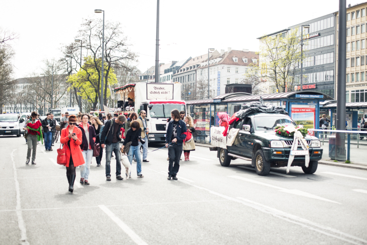 Photograph of a demonstration procession on the multi-lane road at Karlsplatz in Munich city centre. The procession is accompanied by two cars: A black pickup truck, decorated with banners and a large wreath with a mourning ribbon on the bonnet. People sit on the loading area. A second vehicle is a white decorated parade lorry adorned with red fabrics, white and green garlands and paper flowers. On the front of the lorry is a sign with the text "Bruder Schieß nicht!" in old German lettering. The procession participants walk on the street next to the floats.