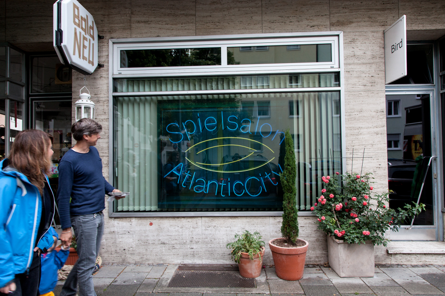 View of a house with a shop with a large window pane. A photograph of the artist Ivan Baschang hangs in the window pane, with a white louvre curtain behind it. The photograph shows a neon sign on a black background. The words "Spielalon Atlantic City" appear on it in blue neon lettering. In front of the window are three planted pots. A man and a woman walk past on the pavement, looking at the window. A child runs between them, holding their hands.