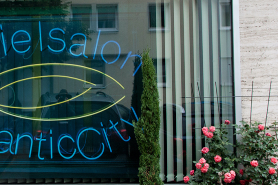 View of a shop window. A photograph by the artist Ivan Baschang hangs in the window pane, behind which is a white louvred curtain. The photograph shows a neon sign on a black background. The words 'Spielalon Atlantic City' are written in blue neon. A flowering rosebush stands at the side of the window.