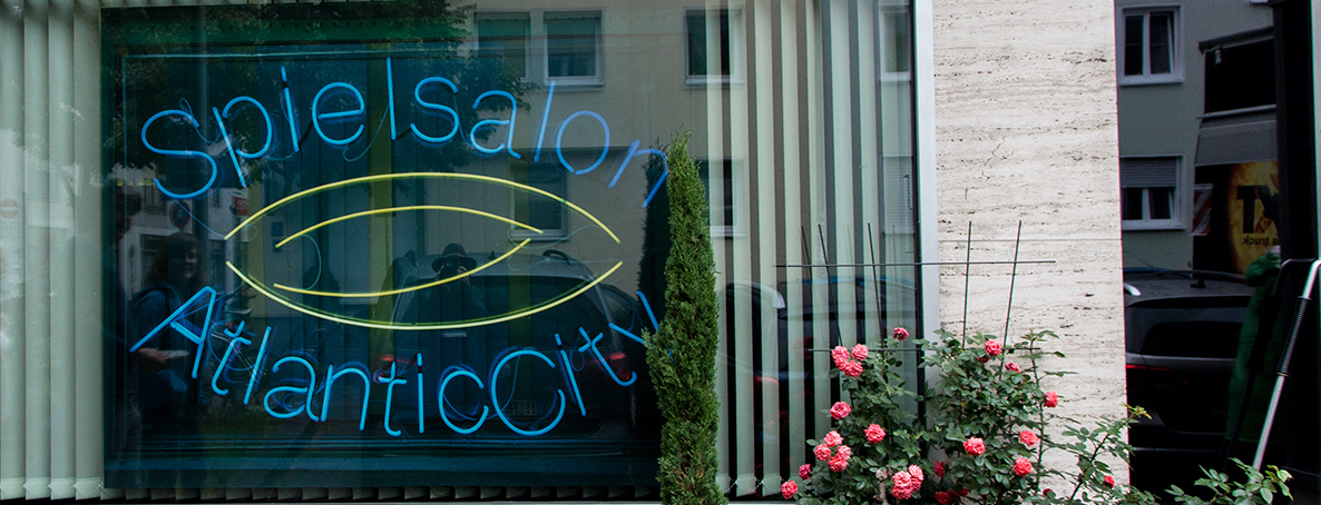 View of a shop window. A photograph by the artist Ivan Baschang hangs in the window pane, behind which is a white louvred curtain. The photograph shows a neon sign on a black background. The words 'Spielalon Atlantic City' are written in blue neon. A flowering rosebush stands at the side of the window.