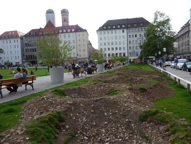 Photograph of an installation by the artist Martin Schmidt. The artist has modelled a crater landscape on an approx. 7 m wide strip of grass on the northern edge of the Marienhof using heaped up and excavated earth. The various craters are slowly overgrown with grass. Benches have been placed on one side of the strip, with plant pots with trees in between. In the background you can see houses on the Weinstraße and the towers of the Frauenkirche.