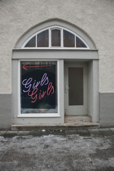 View of a grey façade of a house, in the centre of which is a small shop with a shop window and an entrance door. In the window of the shop is a photograph by the artist Ivan Baschang. The photograph shows a neon sign on a black background. The words 'Girls Girls' and an arrow pointing left appear in pink neon.