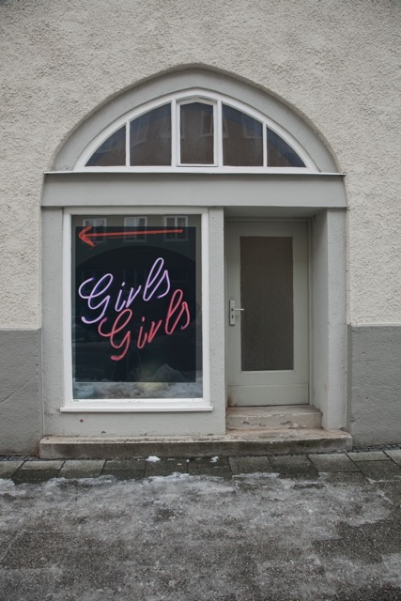 Ansicht einer grauen Hausfassade, im Zentrum zu sehen ein kleines Ladenlokal mit Auslagenfenster und Eingangstür. In der Fensterscheibe des Ladens hängt eine Fotografie des Künstlers Ivan Baschang. Die Fotografie zeigt ein Leuchtreklameschild auf schwarzem Hintergrund. In rosa Leuchtreklameschrift erscheinen dort die Wörter "Girls Girls" sowie ein Richtungspfeil, der nach links zeigt.