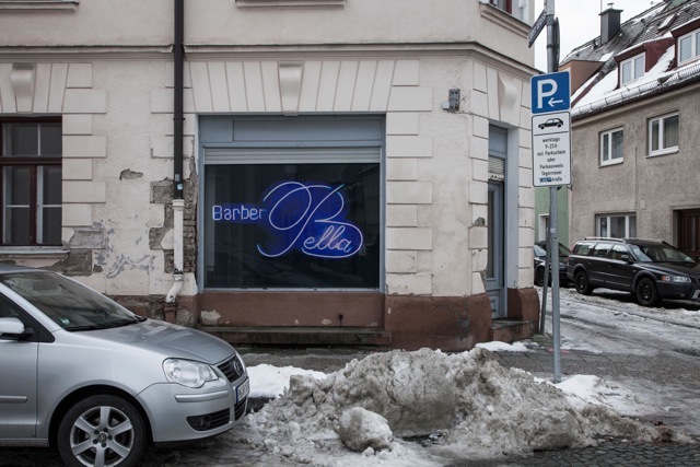 View of the façade of a beige corner building, in the centre a shop with a display window. In the shop window is a photograph by the artist Ivan Baschang. The photograph shows a neon sign on a black background. The words "Barber Bella" are written in blue neon. A car is parked in front of the building and there is slush on the street.