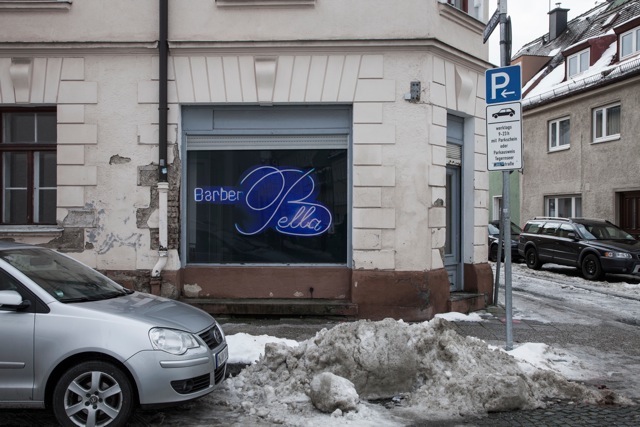 Ansicht der Fassade eines beigen Eckhauses, im Zentrum ein Ladenlokal mit Auslagenfenster. In der Fensterscheibe des Ladens hängt eine Fotografie des Künstlers Ivan Baschang. Die Fotografie zeigt ein Leuchtreklameschild auf schwarzem Hintergrund. Darauf erscheinen in blauer Neonschrift die Wörter "Barber Bella". Vor dem Haus ist ein Auto geparkt und es liegt Schneematsch auf der Straße.