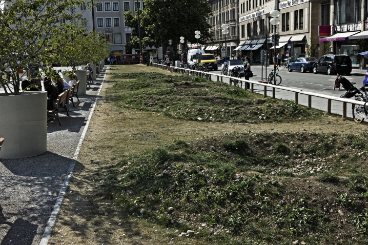 Photograph of an installation by the artist Martin Schmidt. The artist has modelled a crater landscape on an approx. 7 m wide strip of grass on the northern edge of the Marienhof using heaped up and excavated earth. The various craters are slowly overgrown with grass. Benches have been placed on one side of the strip, with plant pots with trees in between.