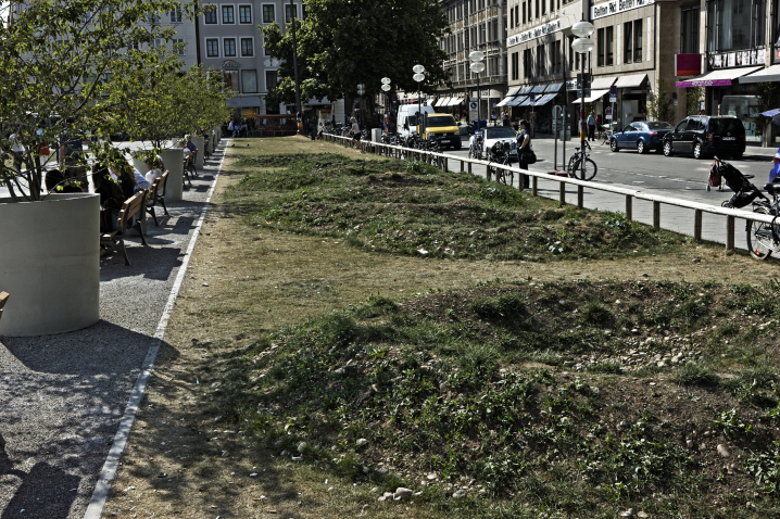 Fotografie einer Installation des Künstlers Martin Schmidt. Auf einem ca. 7 m breiten Rasenstreifen am nördlichen Rand des Marienhofs hat der Künstler aus aufgeschütteter und ausgehobener Erde eine Kraterlandschaft modelliert. Die verschiedenen Krater überwachsen langsam mit Gras. An der einen Seite des Streifens zu sehen sind Bänke, dazwischen stehen Pflanztöpfe mit Bäumen.