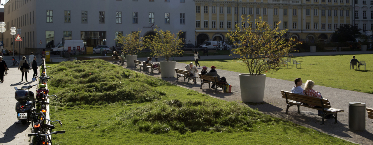 Photograph of an installation by the artist Martin Schmidt. The artist has modelled a crater landscape on an approx. 7 m wide strip of grass on the northern edge of the Marienhof using heaped up and excavated earth. The various craters are overgrown with grass. Benches can be seen on one side of the strip, with plant pots with trees in between. On the other side of the strip, bicycles are leaning against a wooden railing. The houses on Dienerstrasse can be seen in the background.