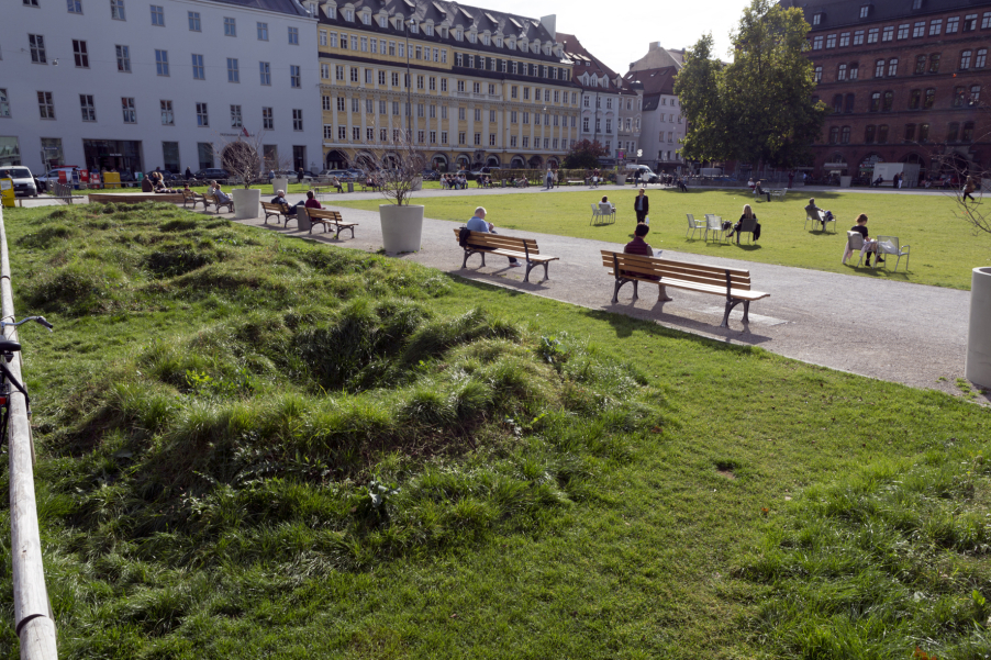Photograph of an installation by the artist Martin Schmidt. The artist has modelled a crater landscape on an approx. 7 m wide strip of grass on the northern edge of the Marienhof using heaped up and excavated earth. The various craters are overgrown with grass. Benches can be seen on one side of the strip, with plant pots with trees in between. On the other side of the strip, bicycles are leaning against a wooden railing. The houses on Dienerstrasse can be seen in the background.