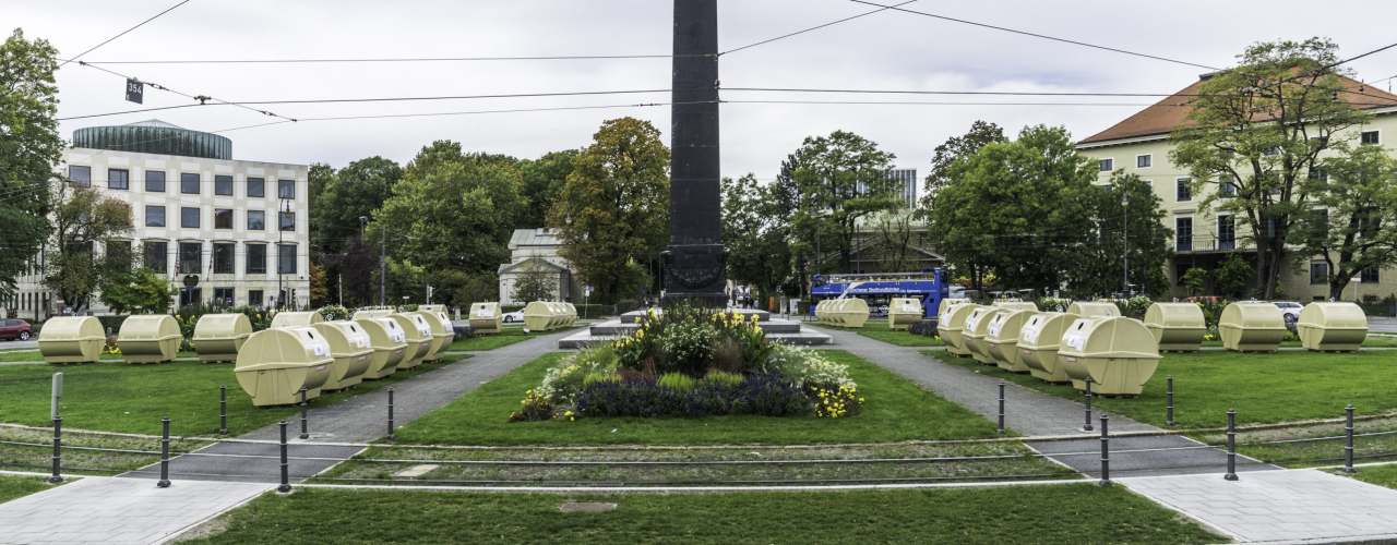 View of Karolinenplatz with the obelisk in the centre and a temporary installation by Lena Bröcker. Rows of pale yellow recycling containers are arranged in a star shape on the green areas of the square.