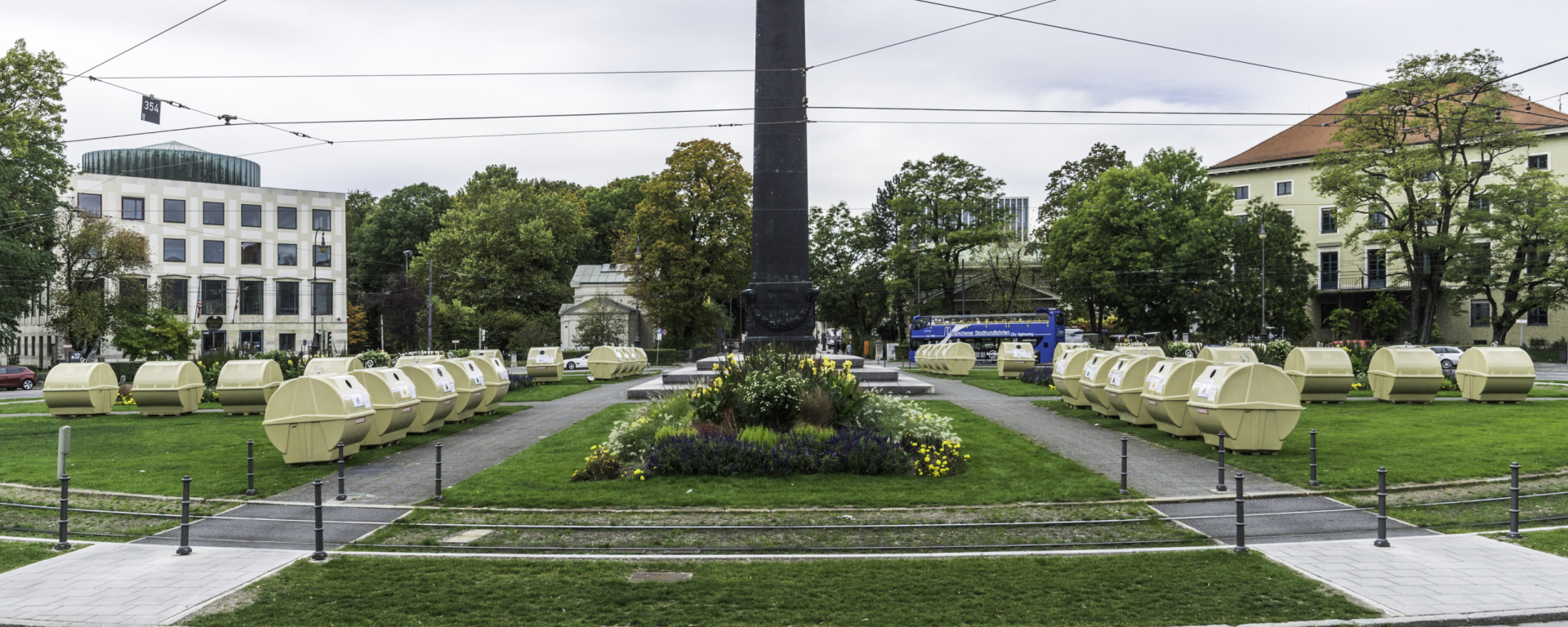 View of Karolinenplatz with the obelisk in the centre and a temporary installation by Lena Bröcker. Rows of pale yellow recycling containers are arranged in a star shape on the green areas of the square.