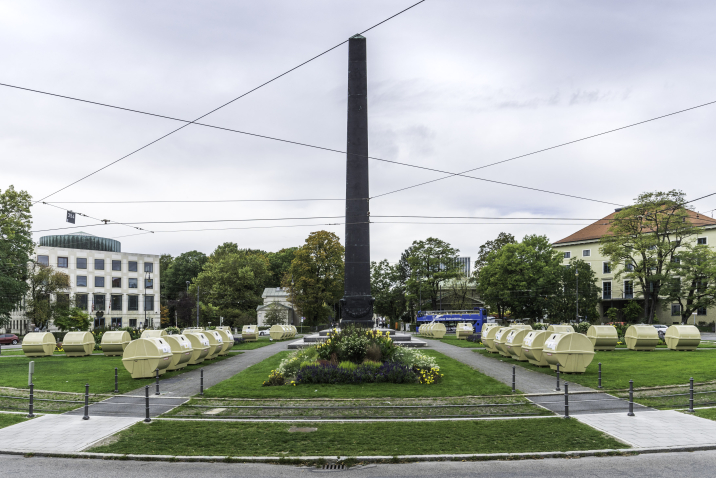 View of Karolinenplatz with the obelisk in the centre and a temporary installation by Lena Bröcker. Rows of pale yellow recycling containers are arranged in a star shape on the green areas of the square.