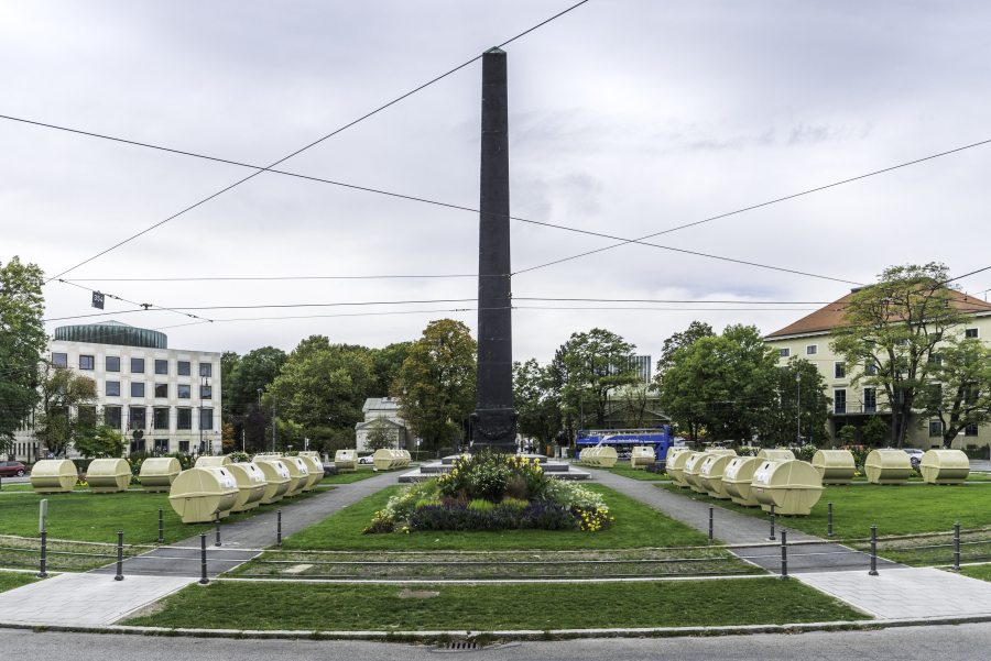 Ansicht des Karolinenplatzes mit dem Obelisken in der Mitte mit einer temporären Installation von Lena Bröcker. Auf den Grünflächen des Platzes sind sternförmig Reihen von blassgelben Wertstoffcontainern aufgestellt.