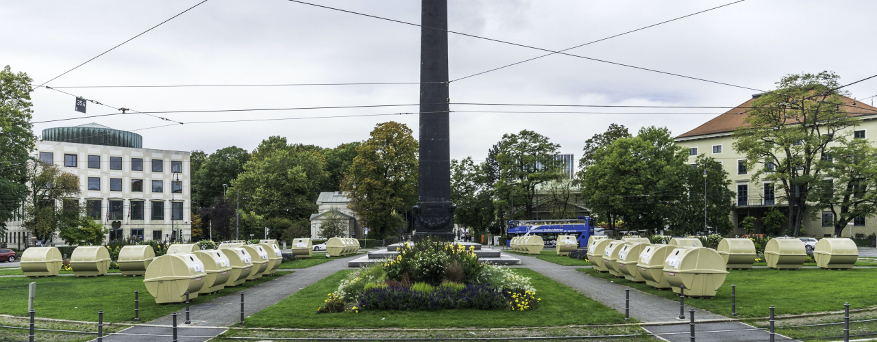 Ansicht des Karolinenplatzes mit dem Obelisken in der Mitte mit einer temporären Installation von Lena Bröcker. Auf den Grünflächen des Platzes sind sternförmig Reihen von blassgelben Wertstoffcontainern aufgestellt.