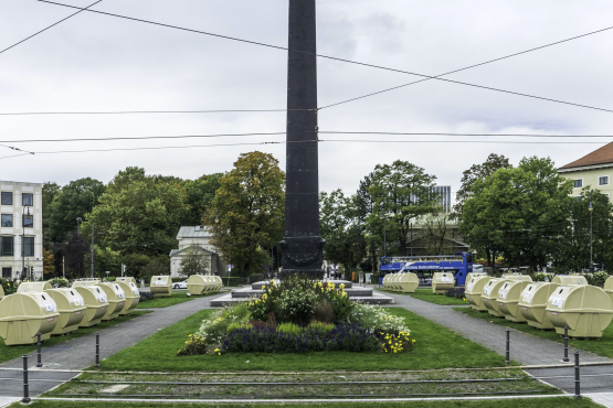 Ansicht des Karolinenplatzes mit dem Obelisken in der Mitte mit einer temporären Installation von Lena Bröcker. Auf den Grünflächen des Platzes sind sternförmig Reihen von blassgelben Wertstoffcontainern aufgestellt.