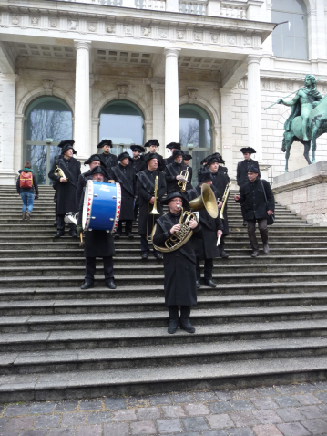 Photograph of a group of men in historical mourning clothing, consisting of a black coat, black trousers and black hats. The men are standing on the steps in front of the entrance to the Academy of Fine Arts, some of them holding musical instruments such as wind instruments and a drum in their hands.