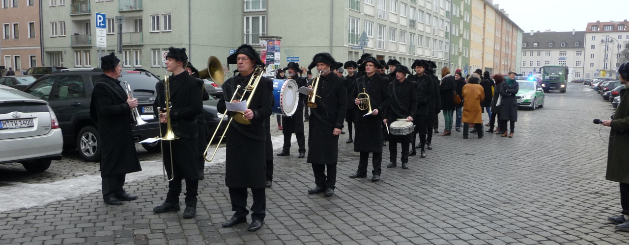 Photograph of a procession on Akademiestraße with residential buildings in the background. Some of the procession participants are dressed in historical mourning clothes, consisting of a black coat, black trousers and black hats. At the front is a band with various instruments playing historical funeral marches during the procession.