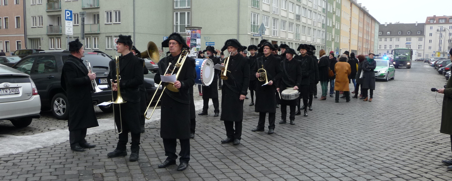 Photograph of a procession on Akademiestraße with residential buildings in the background. Some of the procession participants are dressed in historical mourning clothes, consisting of a black coat, black trousers and black hats. At the front is a band with various instruments playing historical funeral marches during the procession.