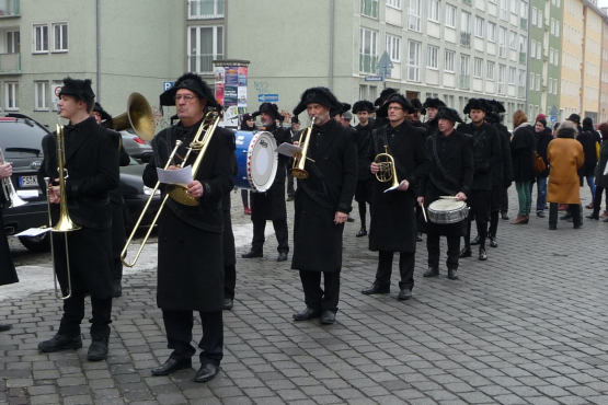 Photograph of a procession on Akademiestraße with residential buildings in the background. Some of the procession participants are dressed in historical mourning clothes, consisting of a black coat, black trousers and black hats. At the front is a band with various instruments playing historical funeral marches during the procession.