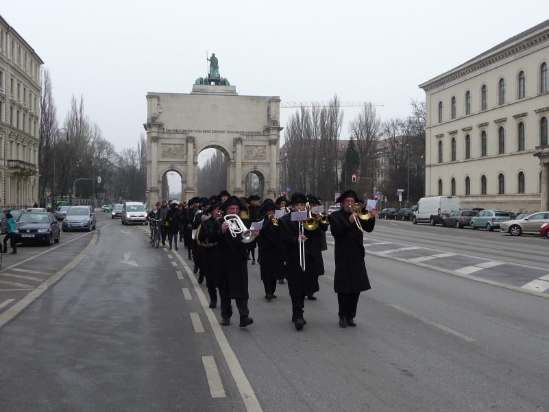 Photograph of a procession on Ludwigstraße with the Siegestor in the background. Some of the procession participants are dressed in historical mourning clothes, consisting of a black coat, black trousers and black hats. A ten-piece band leads the procession, playing historical funeral marches.