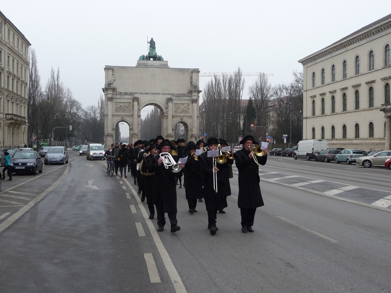 Fotografie einer Prozession auf der Ludwigstraße mit dem Siegestor im Hintergrund. Ein Teil der Prozessionsteilnehmer ist in historische Trauerkleidung gekleidet, bestehend aus schwarzem Mantel, schwarzen Hosen und schwarzen Hüten. An der Spitze geht eine zehnköpfige Kapelle, die während der Umzugsprozession historische Trauermarschmusik spielt.