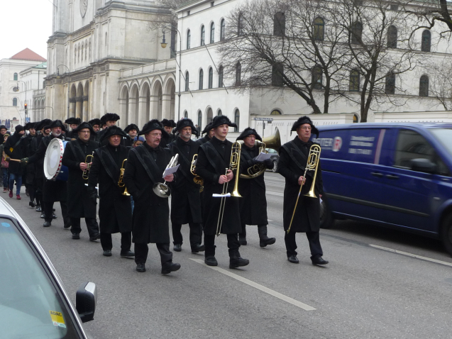 Photograph of a procession on Ludwigstraße. Some of the procession participants are dressed in historical mourning clothes, consisting of a black coat, black trousers and black hats. A ten-piece band leads the procession, playing historical funeral marches.