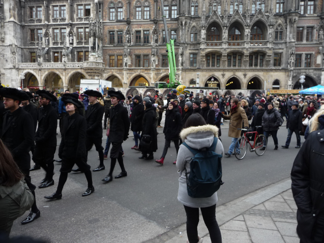 Fotografie einer Prozession auf dem menschengefüllten Marienplatz mit dem Neuen Rathaus im Hintergrund. Ein Teil der Prozessionsteilnehmer*innen ist in historische Trauerkleidung gekleidet, bestehend aus schwarzem Mantel, schwarzen Hosen und schwarzen Hüten.