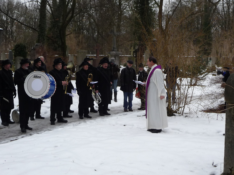 Photograph of a group of people on the snow-covered Old South Cemetery. Some men are dressed in historical mourning clothes, consisting of a black coat, black trousers and a black hat, and are holding musical instruments in their hands. Rainer Maria Schießler, parish priest of St Maximilian's, stands opposite the group and delivers a eulogy.