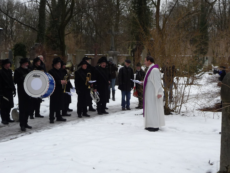 Fotografie einer Gruppe von Menschen auf dem schneebedeckten Alten Südfriedhof. Einige Männer sind in historische Trauerkleidung gekleidet, bestehend aus schwarzem Mantel, schwarzer Hose und schwarzem Hut, und halten Musikinstrumente in der Hand. Rainer Maria Schießler, Pfarrer von St. Maximilian, steht der Gruppe gegenüber und hält eine Grabrede.