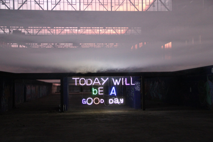 View of the empty industrial hall 24 of the former Freimann railway repair works at sunset. The inside of the hall is already in darkness, in the middle of the hall you can see an installation by artist Emauel Mooner, a colourful neon sign with the words "Today will be a good day". Above it, grey wafts of mist billow from a fog machine.
