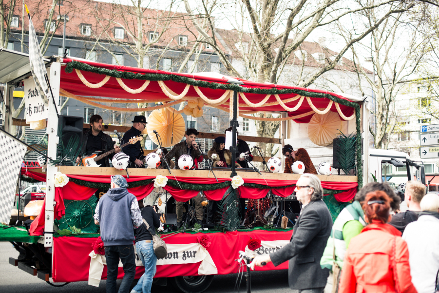 Photograph of a demonstration procession on a street in Munich city centre. The float is decorated with red fabrics, white and green garlands and paper flowers. A band with instruments is on the float, the members are singing into microphones attached to the float.