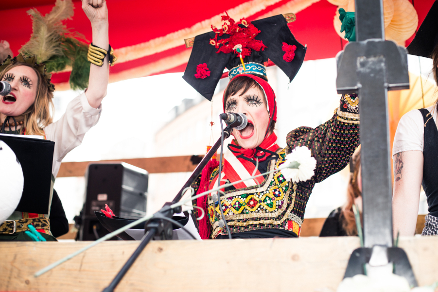 Photo of a float decorated with red fabric, white and green garlands and paper flowers. Several women stand on the float and two sing into microphones attached to the float. They are dressed in colourful traditional costumes, wearing headdresses and expressive make-up.